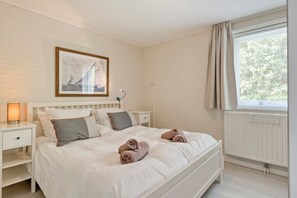 Une chambre à coucher avec un lit, des tables de chevet, une lampe, un tableau encadré et une fenêtre avec des rideaux.