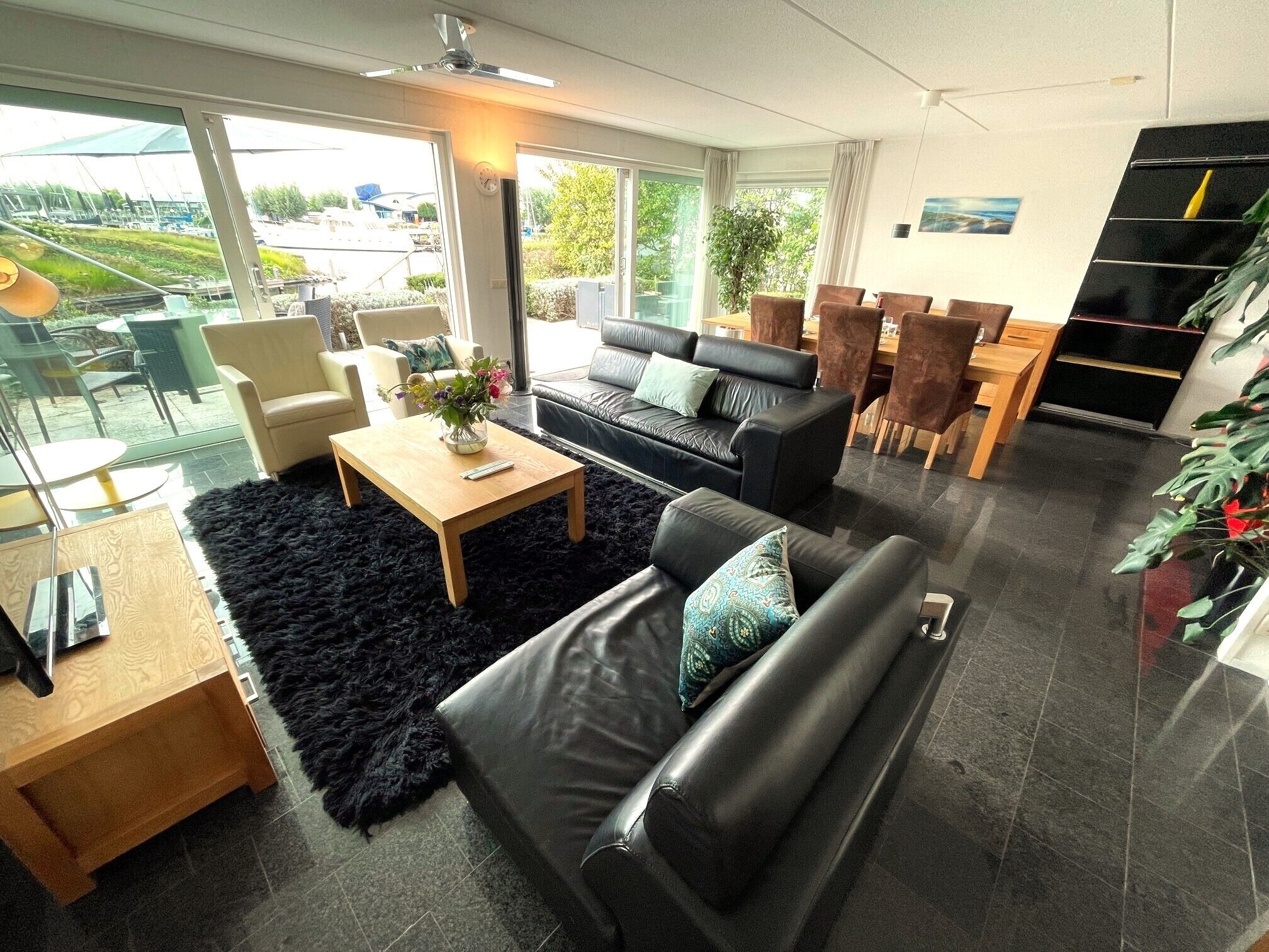 Een moderne woonkamer met een zwarte leren bank, een houten salontafel en een zacht zwart vloerkleed. Er is een eethoek met een houten tafel en stoelen, en een groot raam met uitzicht op de omgeving.