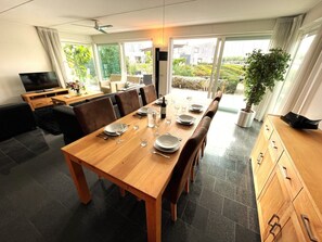 Ein Essbereich mit einem Holztisch, gedeckt für vier Personen, einem Fernseher und Blick auf den Garten durch große Fenster.