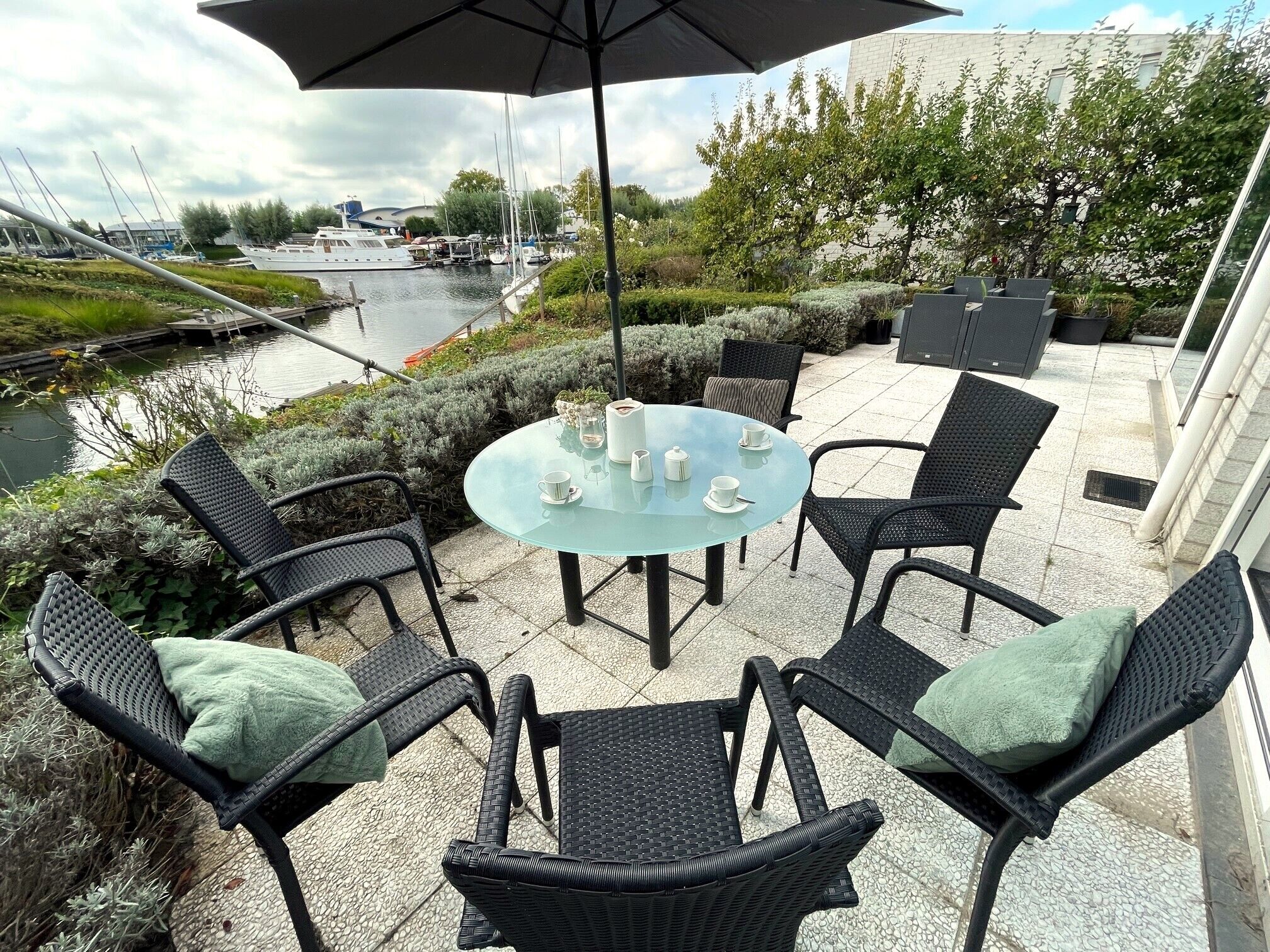Een terras buiten met een ronde glazen tafel, zwarte rieten stoelen en een grote parasol.