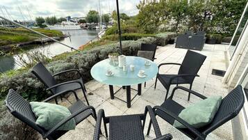 Een terras buiten met een ronde glazen tafel, zwarte rieten stoelen en een grote parasol.