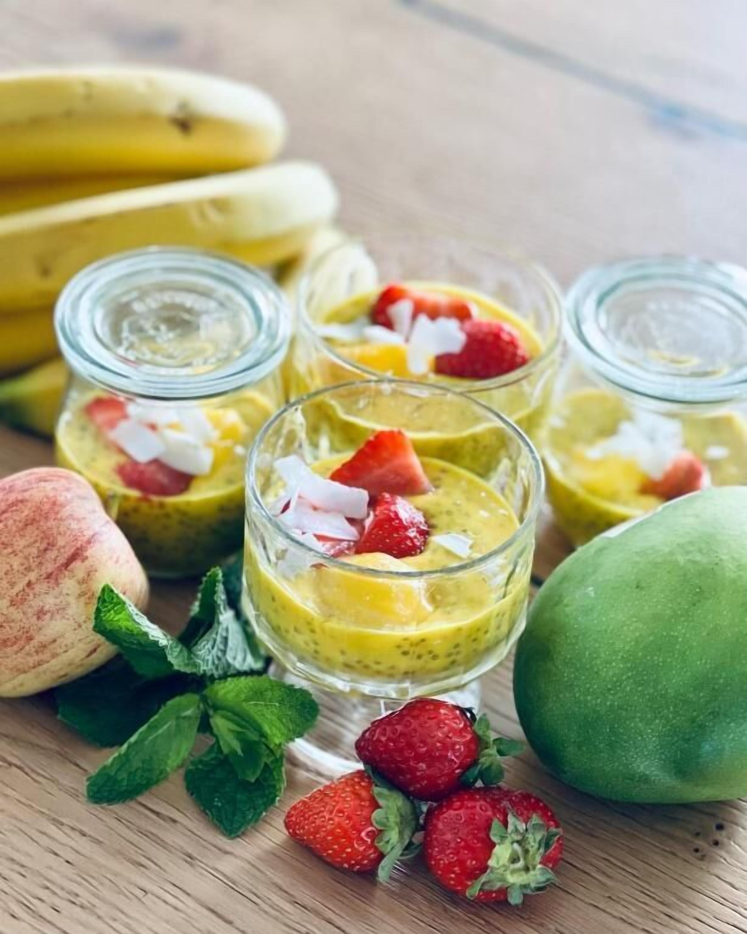 Disposição de sobremesas com pequenos copos contendo um smoothie amarelo, coberto com morangos e coco.