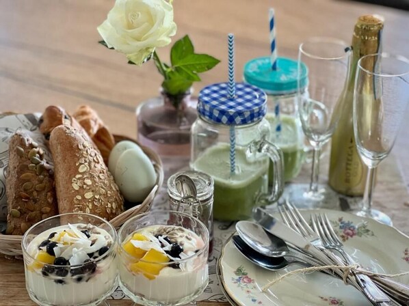 Une table dressée avec du pain, un smoothie vert, deux verres de yaourt aux fruits, une bouteille de champagne et un vase contenant une rose blanche.