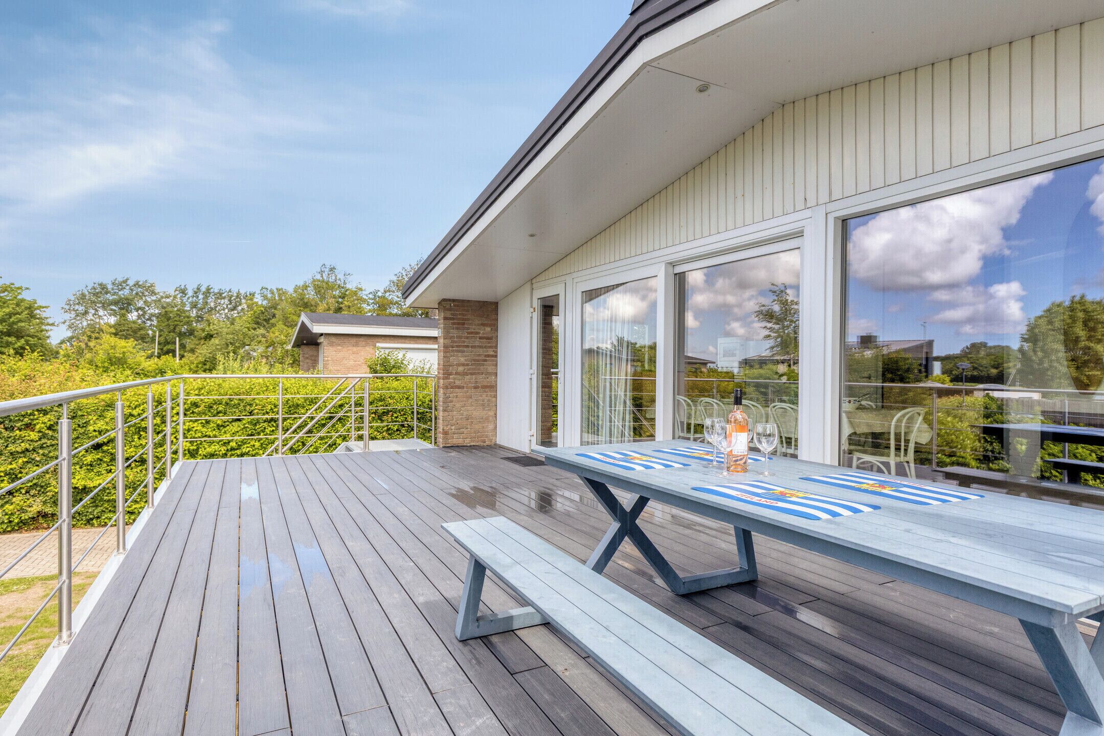 Eine Holzterrasse mit Tisch und Bänken, eine Glastür und Blick auf Grünflächen sowie ein Gebäude im Hintergrund.