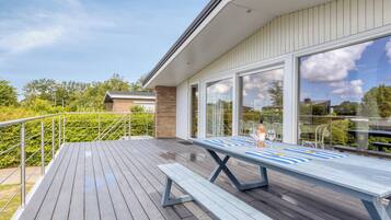 Eine Holzterrasse mit Tisch und Bänken, eine Glastür und Blick auf Grünflächen sowie ein Gebäude im Hintergrund.