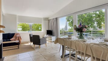 Een moderne woonkamer met een bank, televisie, eettafel en stoelen, grote ramen met uitzicht op groen.