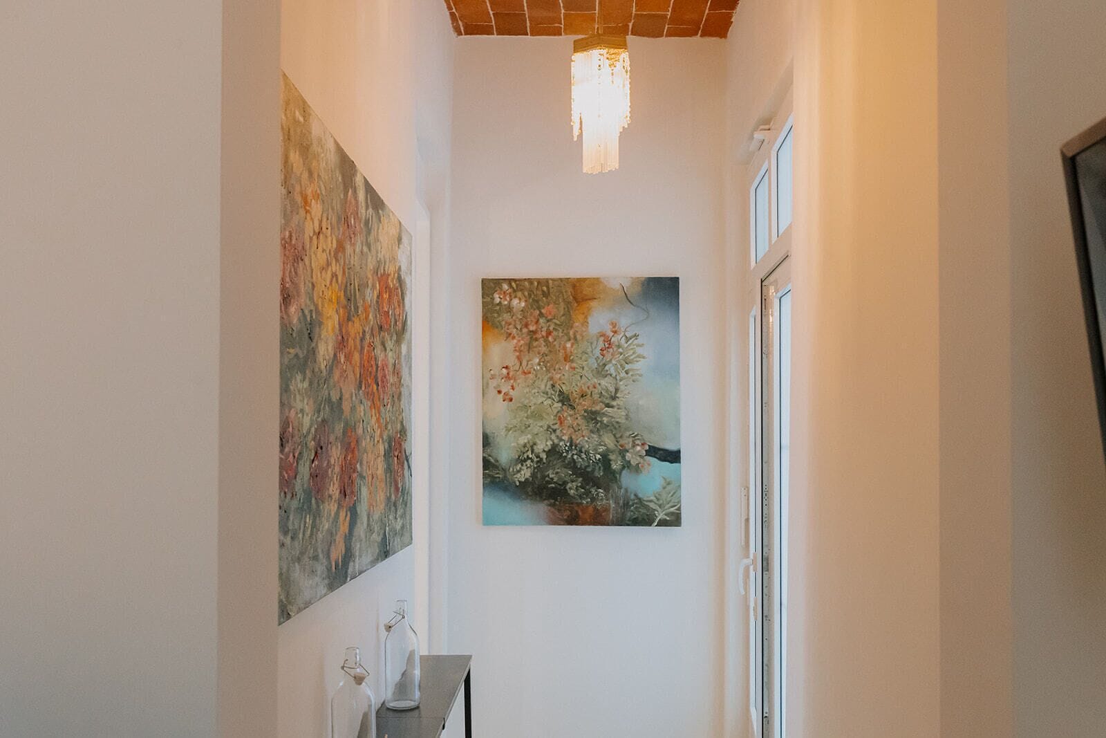 A hallway with two paintings on the walls, a chandelier, and a window on the right.