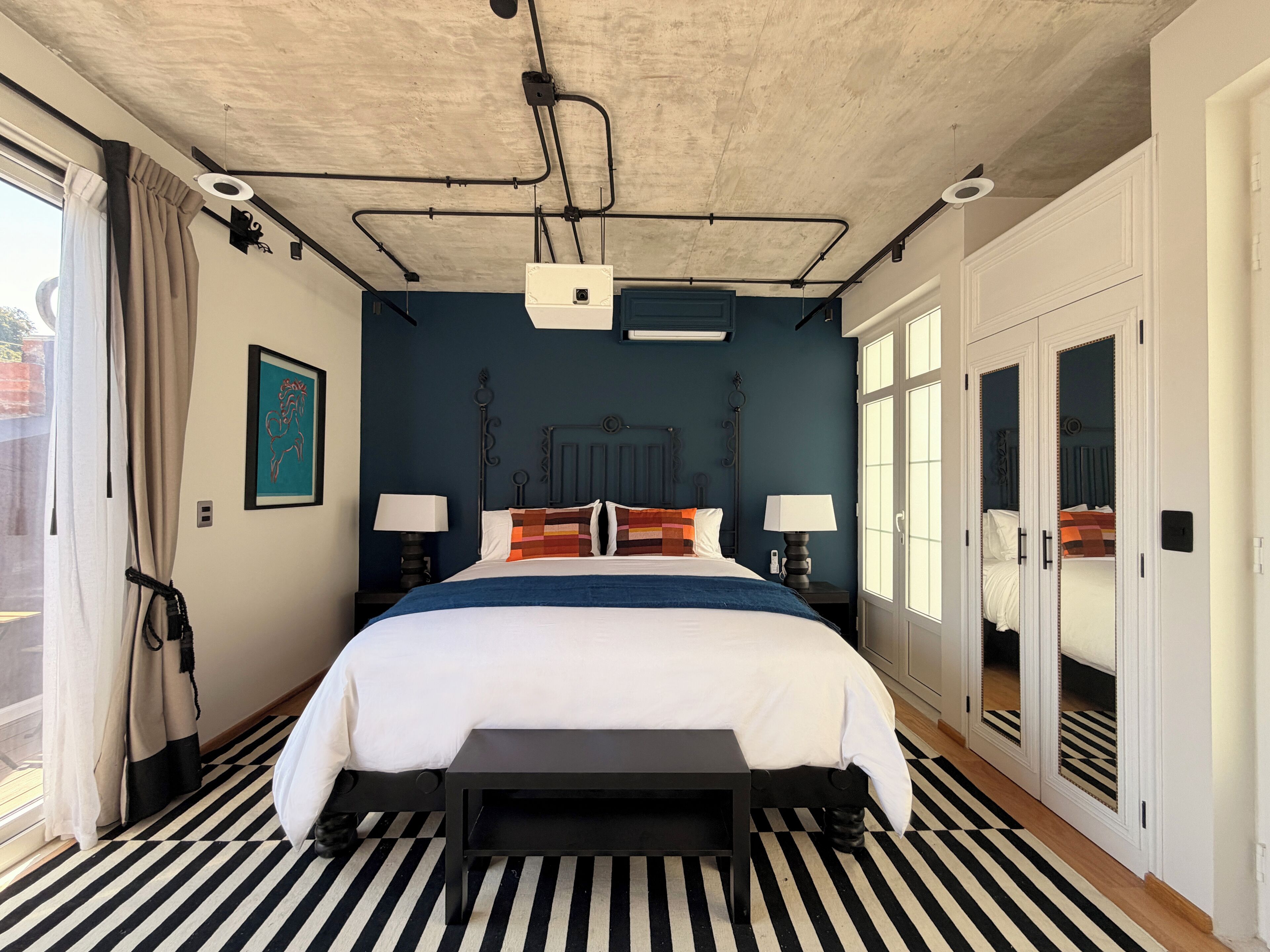 Ein Schlafzimmer mit Bett, Nachttischen, einer Bank, einem Deckenprojektor und einem großen Fenster.