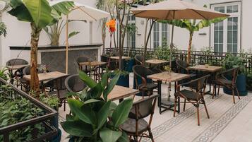 Un patio al aire libre con varias mesas y sillas, rodeado de plantas en macetas y sombrillas.