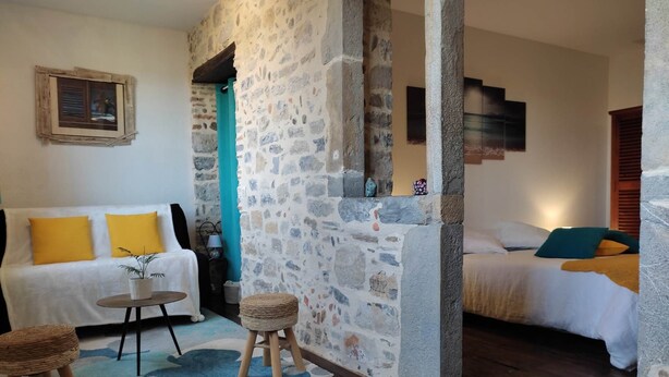 A cozy room with a stone wall, a wooden floor, a white sofa with yellow cushions, a small table with a plant, and a bed with a blue pillow. - Authentic Suite - PATRIMOINE (OLORON STE MARIE)