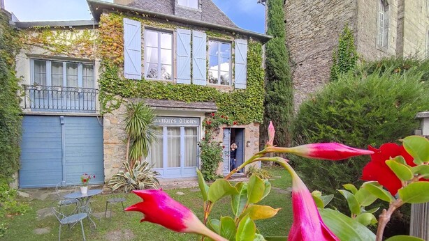 A stone building with a blue garage door, a sign reading "auberges d'hôtes," and a garden with red flowers and a metal chair. - Colorful Room - ASPE (OLORON STE MARIE)