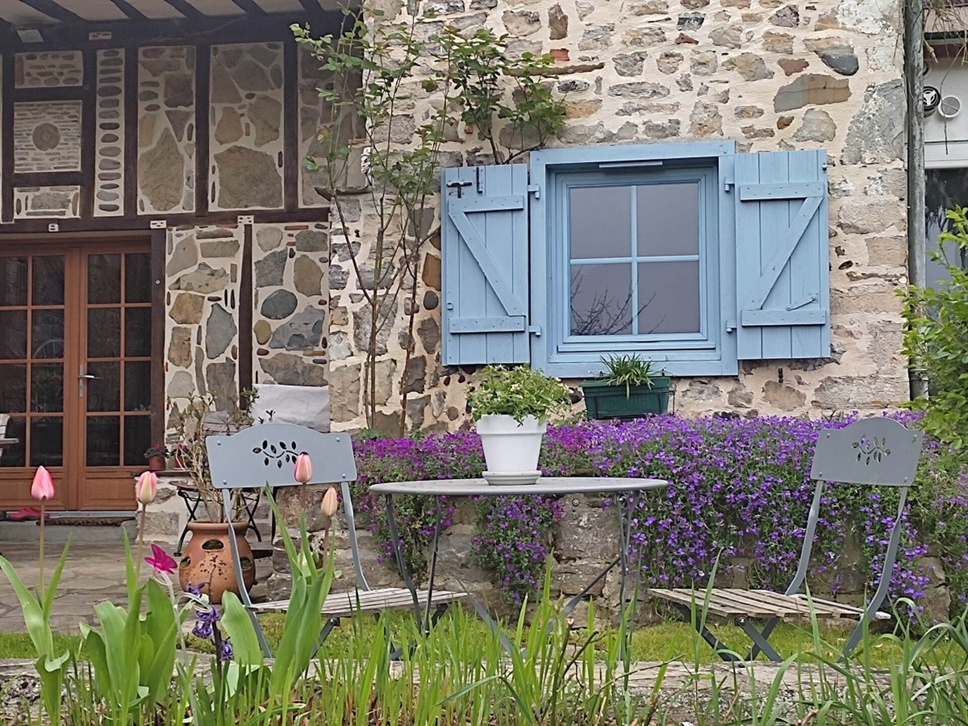 Ein Steinhaus mit einer Holztür und blauen Fensterläden, ein kleiner Tisch mit einem Topfpflanzen und lila Blumen im Hintergrund.