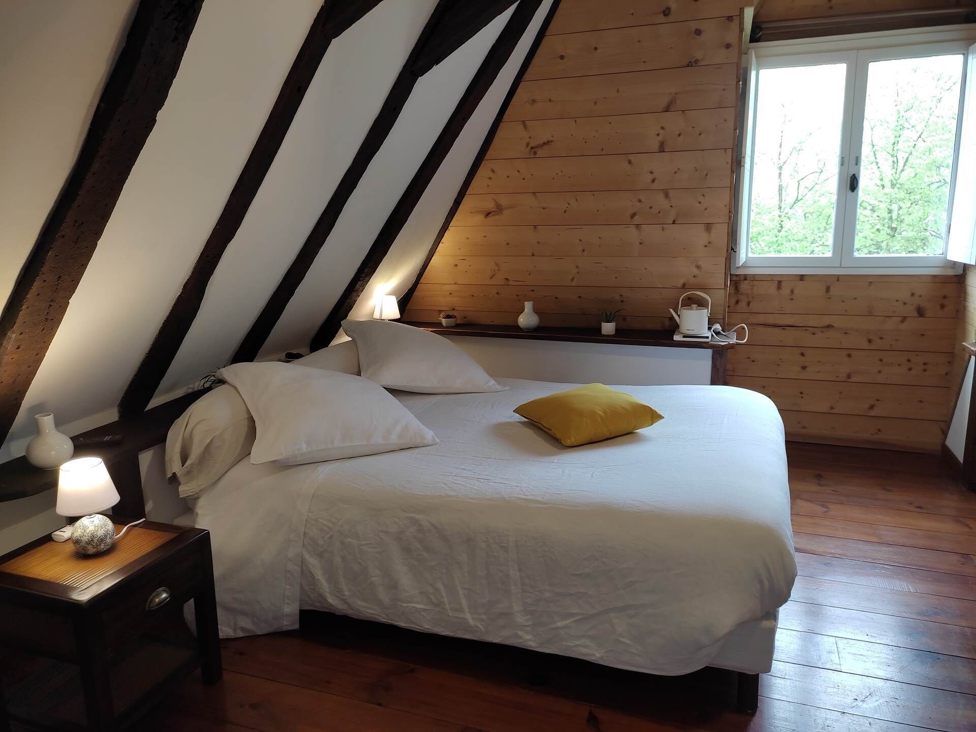 Ein Schlafzimmer mit schrägen Wänden, Holzwänden, einem Bett mit weißer Bettwäsche und einem gelben Kissen, einem Nachttisch mit Lampe sowie einem Fenster mit Blick auf Bäume.