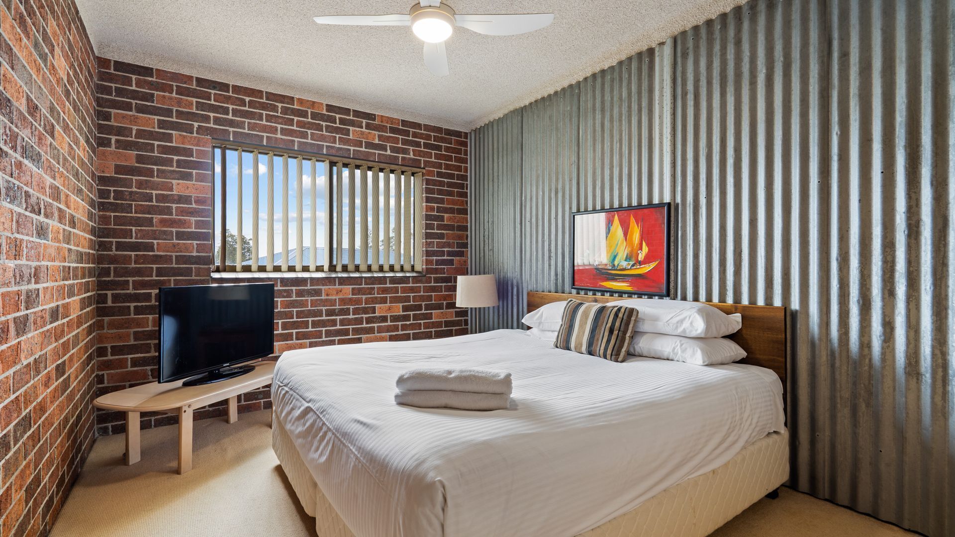 A hotel room with a brick wall, a bed with white bedding, a TV on a wooden stand, a window with blinds, a painting on the wall, and a ceiling fan.