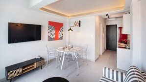 Un salon moderne comprenant un coin repas, un téléviseur fixé au mur et une cuisine visible à travers une porte ouverte.