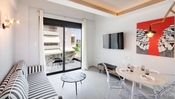 Un salon moderne avec un canapé à rayures, une table de salle à manger ronde entourée de chaises, un téléviseur à écran plat fixé au mur et une grande fenêtre donnant sur l’extérieur.