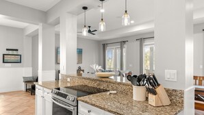 Cozinha moderna com bancadas de granito, armários brancos e uma ilha grande. Há luminárias pendentes, um ventilador de teto e uma janela com persianas.