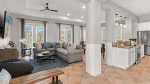 Ampla sala de estar com sofá seccional cinza, mesa de centro de madeira e espaço de jantar com mesa pequena e cadeiras. Cozinha visível, com armários brancos e eletrodomésticos de aço inoxidável.