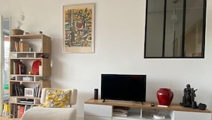 Una sala de estar con un televisor de pantalla plana, un sofá blanco, un tapete estampado, una estantería y un cuadro en la pared.