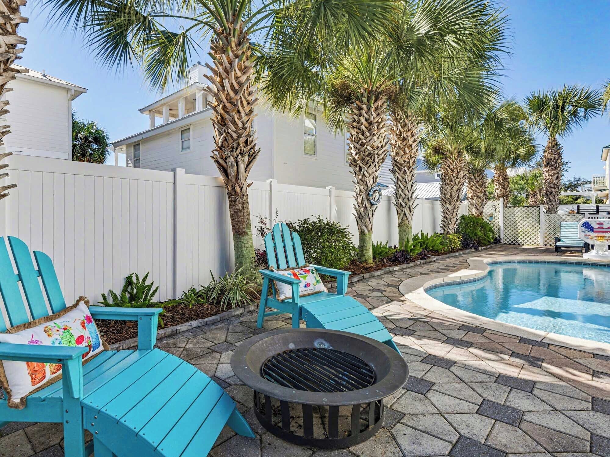 A pool area with a fire pit, two turquoise Adirondack chairs, and a white fence.