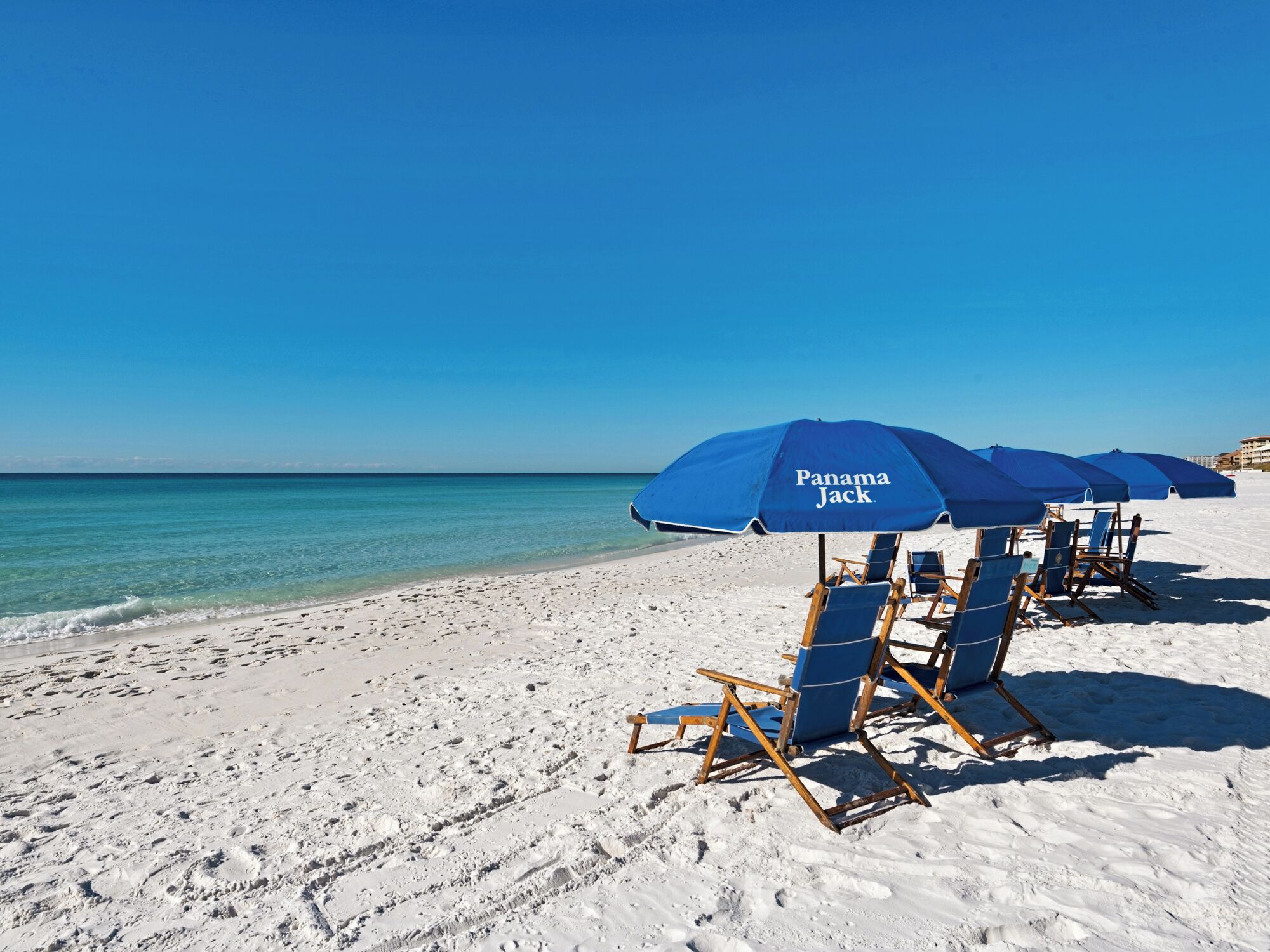 A beach with white sand and clear blue water. There are blue umbrellas and wooden beach chairs arranged on the sand.