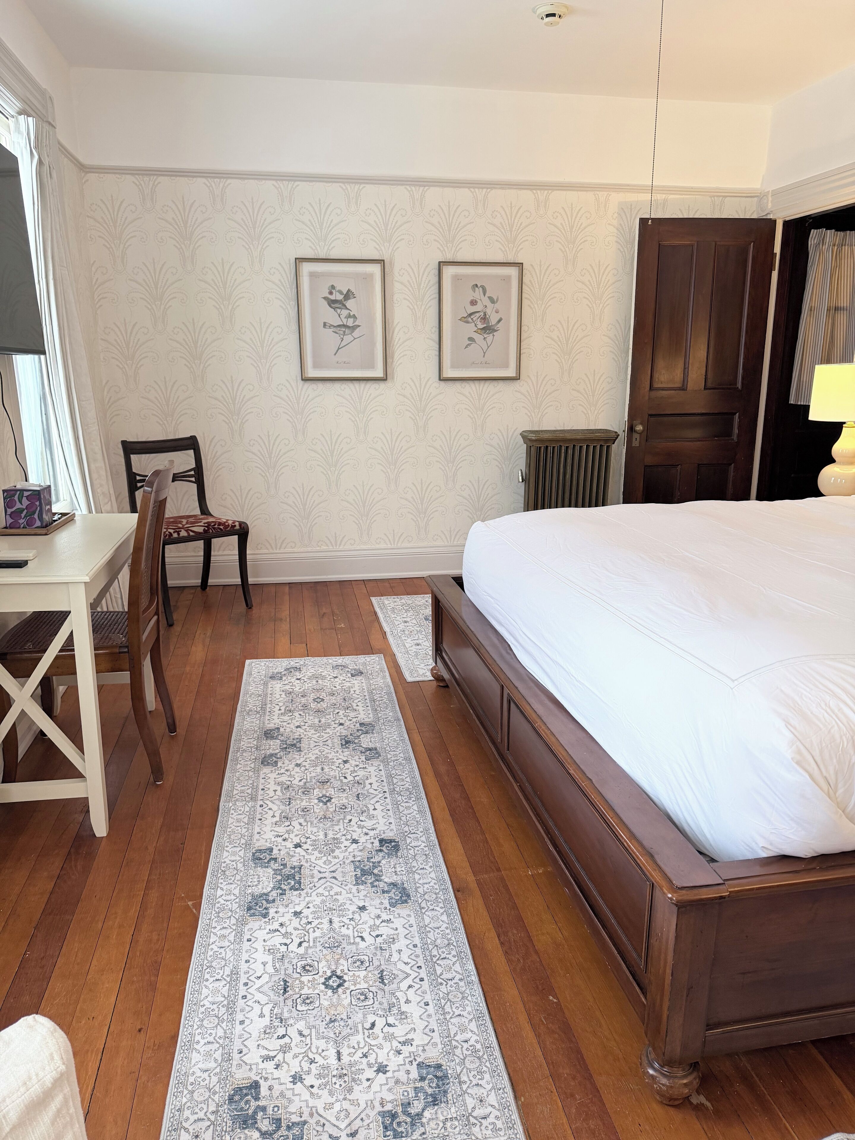 A bedroom with a large bed, a wooden desk, a chair, and two framed botanical prints on the wall.