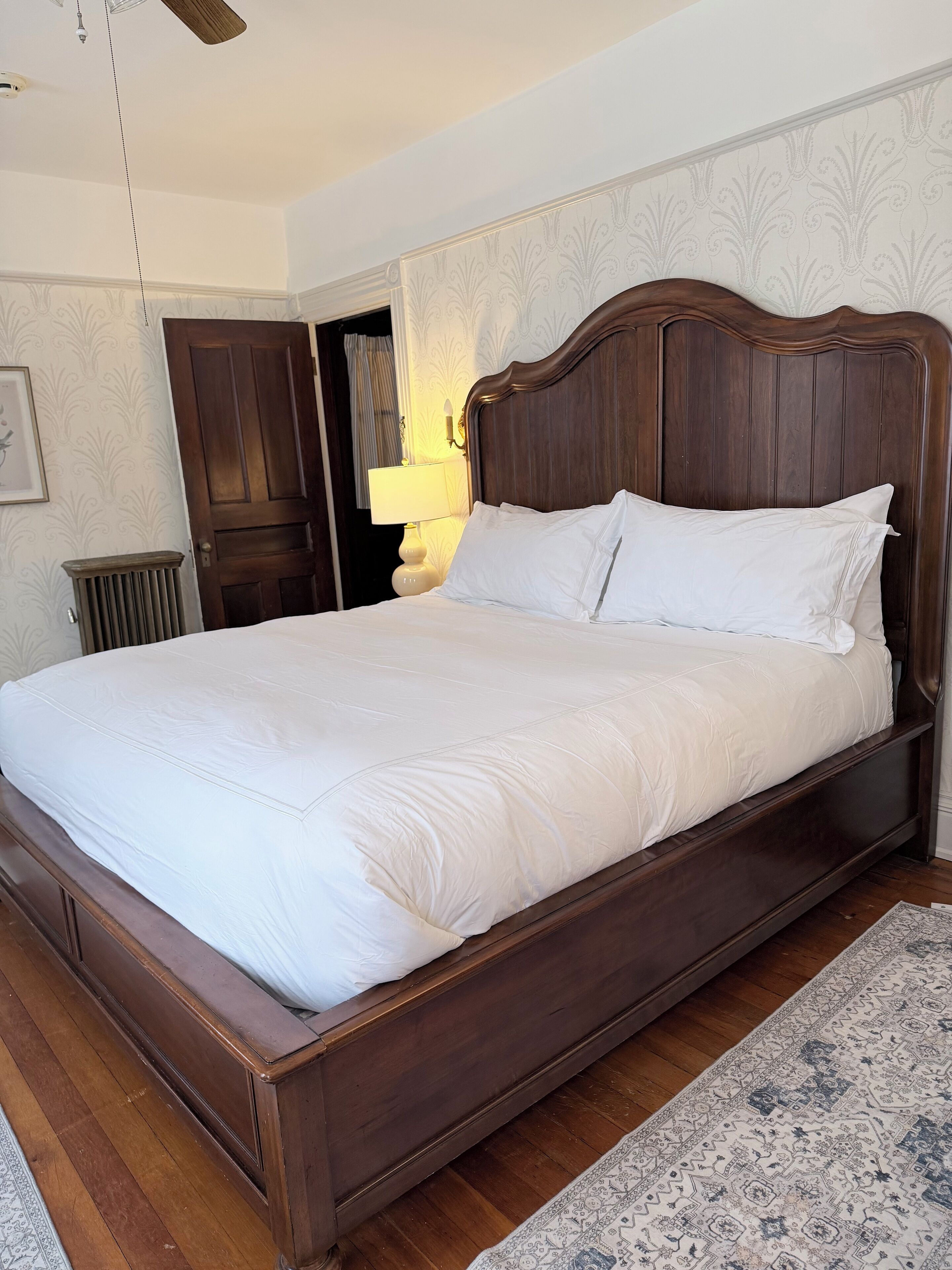 A large, dark wooden bed with white bedding in a room with patterned wallpaper and a wooden floor.