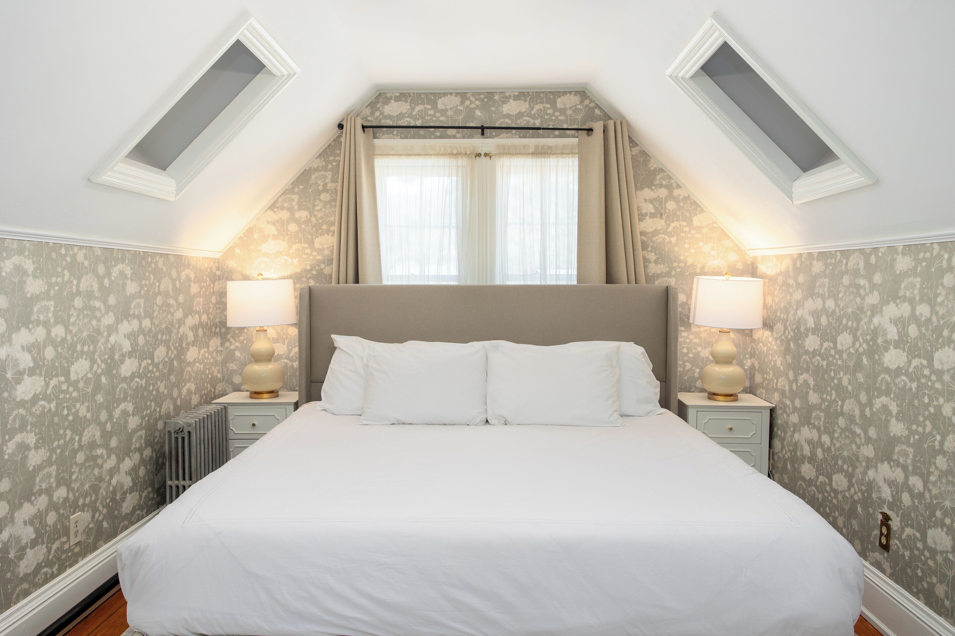 A bedroom with a large bed, two bedside lamps, a radiator, and a window with curtains.