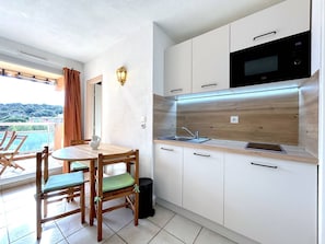 Eine kleine Küche mit Mikrowelle, Spüle und Holzschränken. Es gibt einen Esstisch mit Stühlen und einen Blick auf einen Balkon.