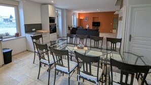 Una cocina moderna con armarios blancos, horno empotrado y microondas. Hay una zona de comedor con mesa y sillas de vidrio.
