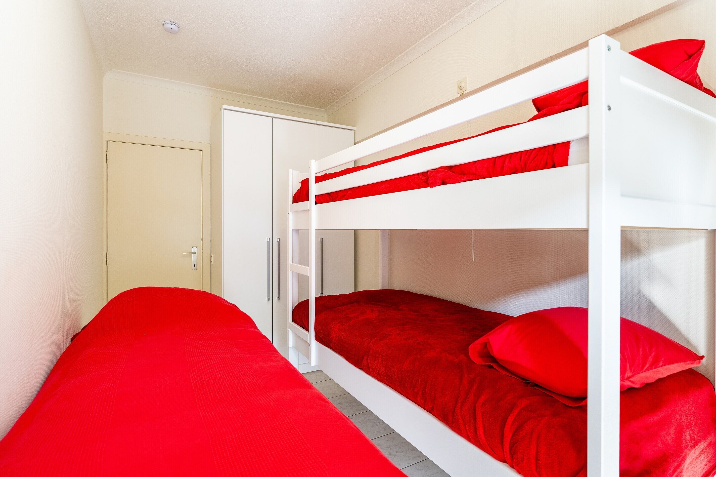 Una cama individual con ropa de cama roja en una habitación con paredes blancas y una puerta cerrada.