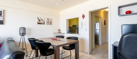 A modern living room with a dining area, a kitchen in the background, and a small office space.