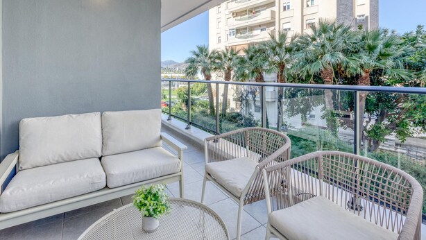 A balcony with a sofa and wicker chairs, overlooking a residential area with palm trees and a building in the background. - Modern apartment in Estepona with parking, near beaches (Estepona)