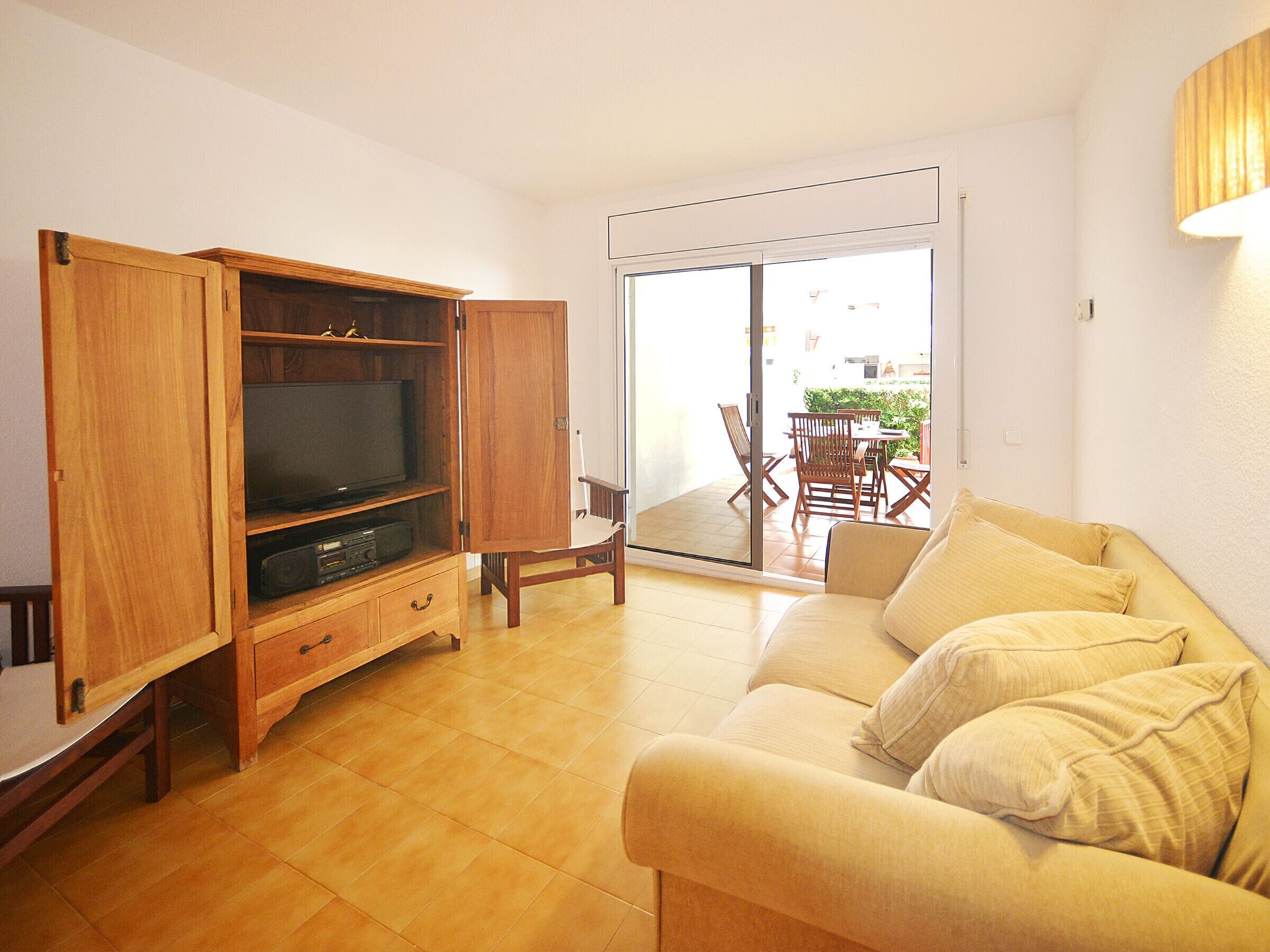 A living room with a wooden entertainment unit, a flat-screen TV, a beige sofa, and a sliding glass door leading to an outdoor patio with a table and chairs.