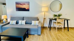 Ett modernt vardagsrum med en grå soffa, ett soffbord, ett skrivbord med en stol, en golvlampa, en rund spegel och en inramad tavla som föreställer en solnedgång.