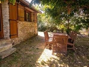 Un bâtiment en pierre avec des volets en bois, une table en bois avec des chaises et un arbre qui offre de l’ombre.