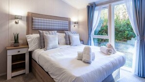 A neatly made bed with white linens and a plaid headboard, a bedside lamp, and a view of greenery outside. - Azenor Lodge - Kelling Heath Holiday Park - 16221 (Holt)