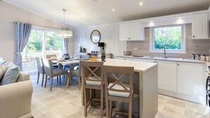 A modern kitchen with a dining area, featuring a central island, white cabinetry, and a large window. - Azenor Lodge - Kelling Heath Holiday Park - 16221 (Holt)