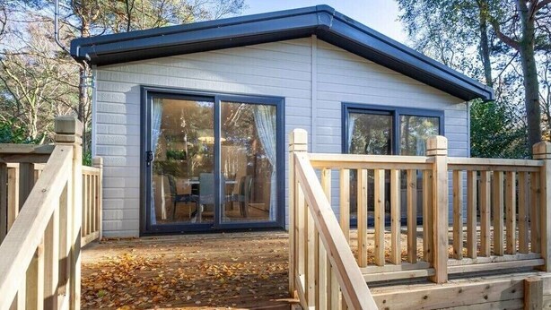 A small, single-story cabin with a deck and a sliding glass door leading to a patio area. - Azenor Lodge - Kelling Heath Holiday Park - 16221 (Holt)