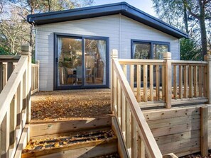 A small, single-story cabin with a deck and a sliding glass door leading to a patio area. - Azenor Lodge - Kelling Heath Holiday Park - 16221 (Holt)