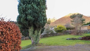 Un camí de grava que condueix a un mur de pedra, un gran arbre de fulles perennes i una zona verda amb un mur de pedra i un turó al fons.
