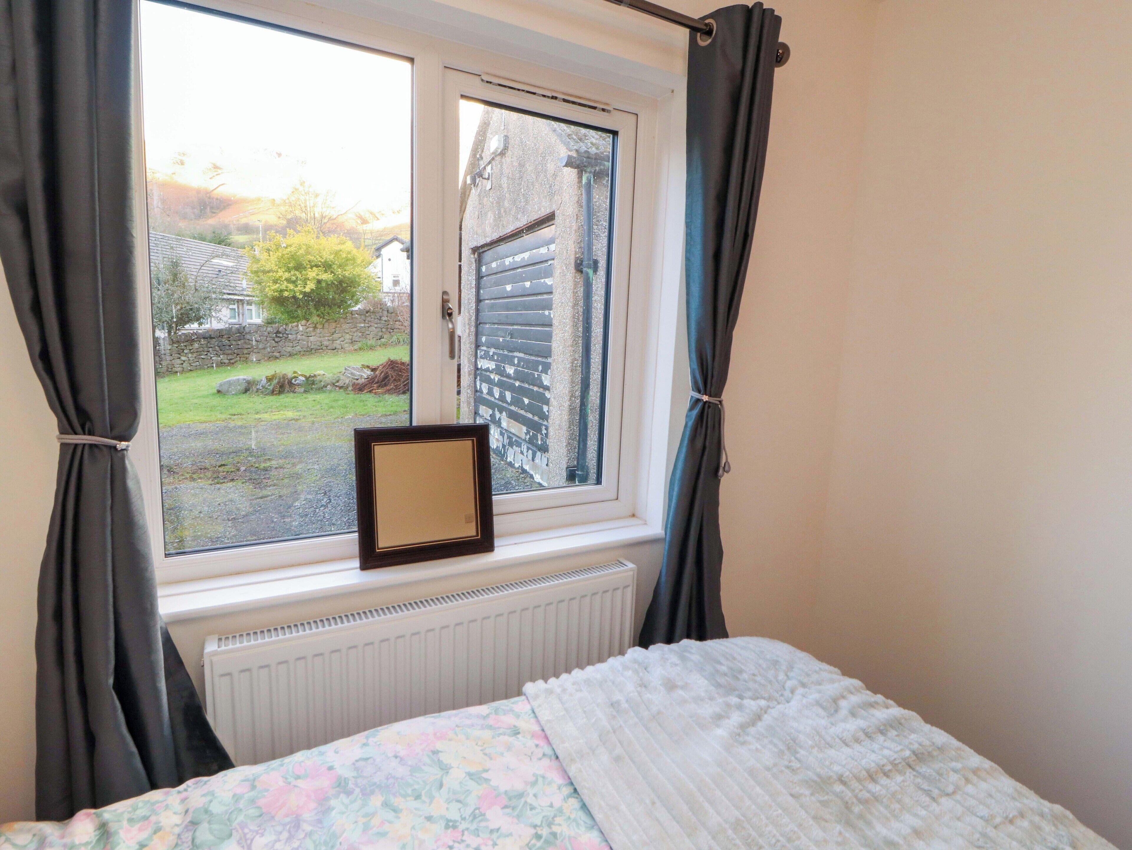 A bedroom with a bed, a window with curtains, a radiator, and a view of a garden and buildings outside.