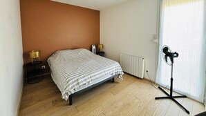Un dormitorio con cama, lámparas de noche, radiador, ventilador y ventana con cortinas.