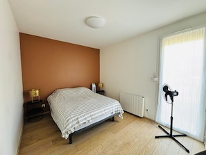 Ein Schlafzimmer mit Bett, Nachttischlampen, Heizkörper, Standventilator und einem Fenster mit Vorhängen.