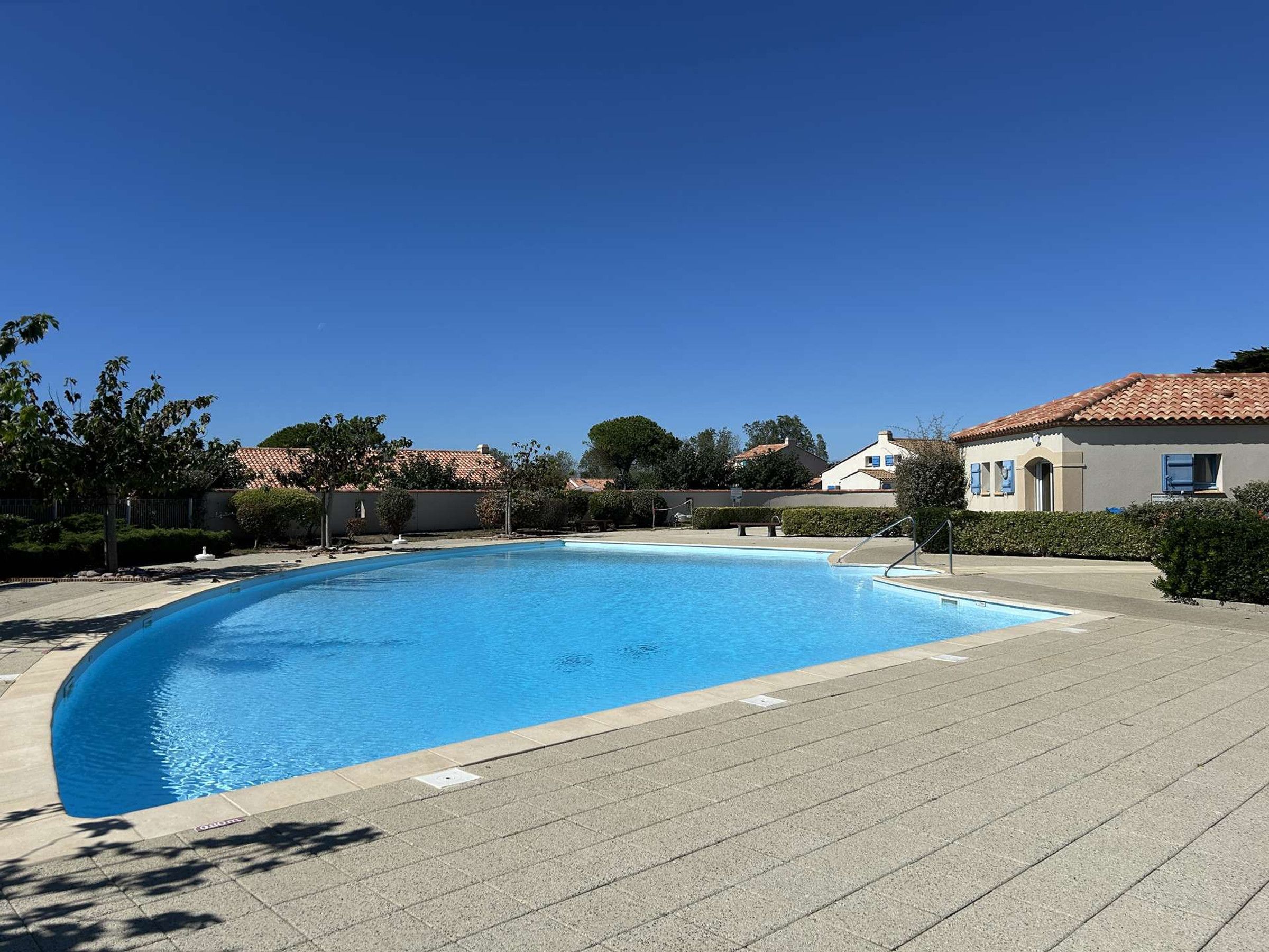 Une piscine à bord incurvé, entourée d’une zone pavée agrémentée de quelques arbres et d’un bâtiment doté d’un toit en tuiles.