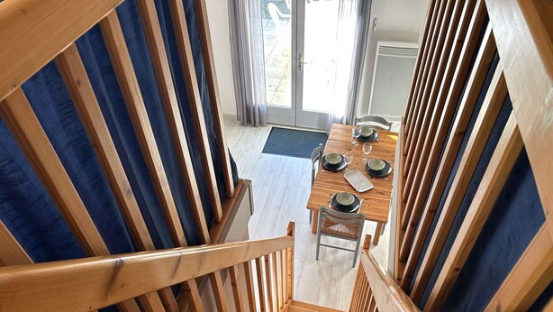 Eine Holztreppe führt zu einem Essbereich mit einem Tisch für vier Personen, einem Stuhl und Blick ins Freie durch eine Glastür.