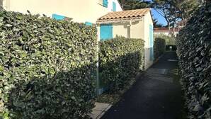 Ein schmaler Weg, gesäumt von ordentlich geschnittenen Hecken, der zu einem Gebäude mit Ziegeldach und blauen Fensterläden führt.