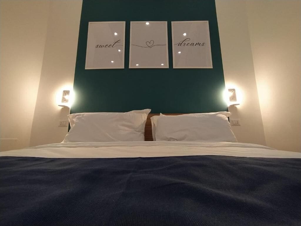 A neatly made bed with white linens and a dark blue blanket, flanked by two wall-mounted light fixtures. Above the bed, there are three framed signs with the words "sweet," "dreams," and a heart symbol.