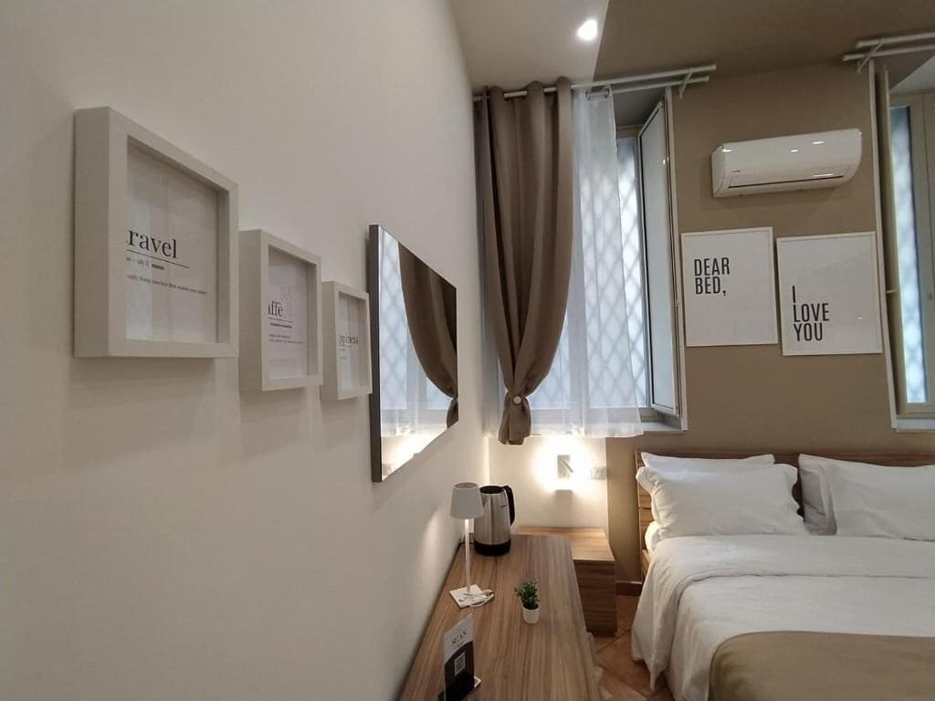 A modern hotel room with a bed, a bedside table with a lamp, a small plant, and framed pictures on the wall.