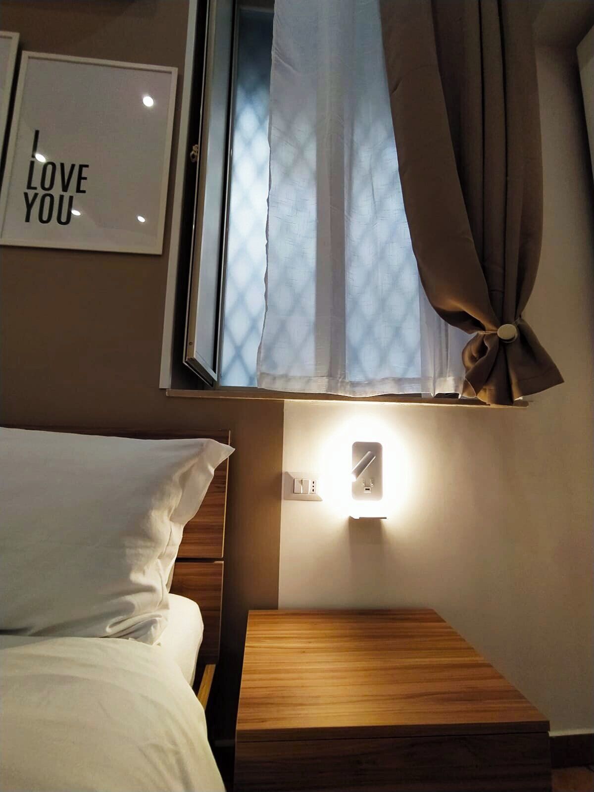 A hotel room with a bed, a bedside table, a wall-mounted light, a window with curtains, and a framed sign that says "I LOVE YOU".